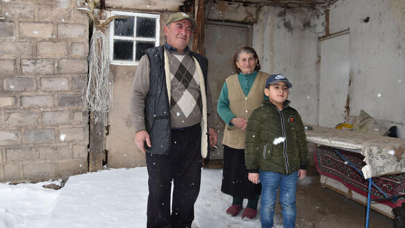Ein alter Mann, eine alte Frau und ein kleiner Junge stehen unter einem Vordach eines Hauses, es fällt Schnee.