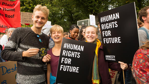 Drei junge Menschen stehen inmitten einer großen Menschenmenge und lächeln in die Kamera. Zwei von ihnen halten Plakate hoch, auf denen steht: "Human Rights for Future".
