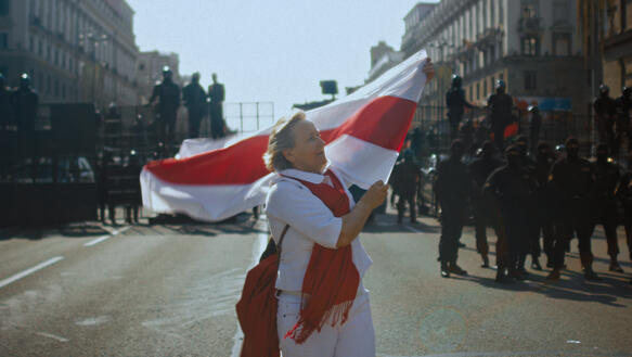 Eine Frau schwingt die belarussische Flagge bei einer Demonstration vor Panzern und Sicherheitskräften.