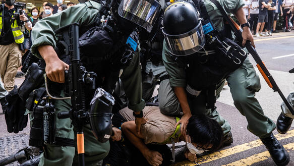 Zwei uniformierte und bewaffnete Sicherheitskräfte drücken einen Demonstranten zu Boden.