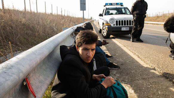 Zwei junge Männer sitzen am Straßenrand. Im Hintergrund steht ein uniformierter Polizist und ein Polizeiwagen.