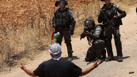 Das Bild zeigt einen Mann mit erhobenen Armen und einer palästinensischen Flagge. Vor ihm befinden sich drei Soldaten der israelischen Grenzpolizei, ein Beamter zielt auf ihn.