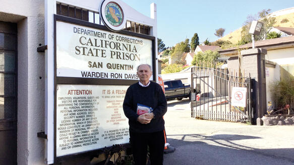 Ein älterer Mann, der die Hände vor dem Bauch faltet und eine ZipLock-Bag festhält, steht vor einer Toreinfahrt mit geöffnetem Tor, auf einem Schild steht "California State Prison", im Hintergrund grüne Hügel und Bäume.
