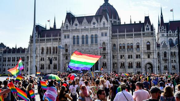 Das Foto zeigt eine große Menschenmenge auf dem Platz vor dem Parlamentsgebäude. Einige Personen schwenken die Regenbogenfahne.