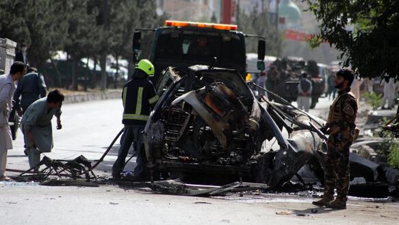 Das Foto zeigt ein ausgebranntes Fahrzeug auf einer Straße. Hilfskräfte haben sich um den Ort des Anschlags versammelt.
