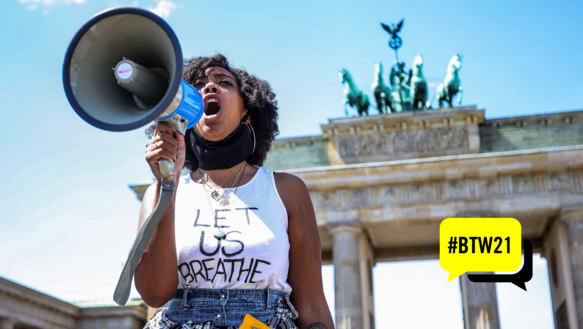 Ein Frau ruft etwas in ein Megaphon. Auf ihrem weißen Shirt hat sie in schwarzer Schrift stehen: "Let us breathe". Im Hintergrund das Brandenburger Tor. In der Bildecke rechts unten eine gelbe Sprechblase, in der mit schwarzer Schrift steht: #BTW21. 