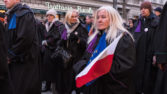 Das Bild zeigt eine Frau mit zwei Flaggen, EU und Polen. Sie demonstriert mit anderen und trägt eine Robe, wie sie Richter_innen tragen