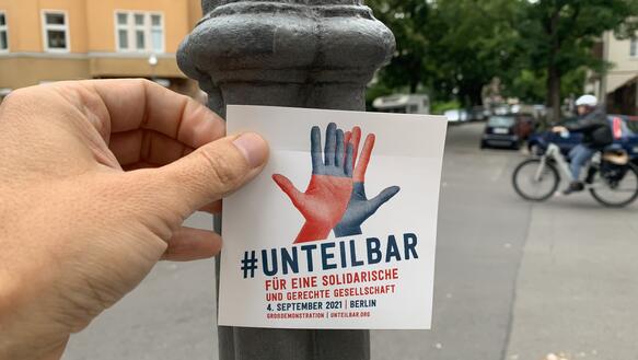 Eine Hand hält einen Sticker vor einen Laternenpfahl. Auf dem Sticker sind zwei Hände abgebildet. Auf dem steht unter anderem: "Unteilbar. 4. September 2021. Großdemonstration". 