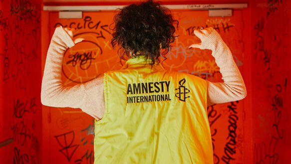 Das Bild zeigt eine Person von hinten mit einer gelben Jacke und Amnesty-Logo