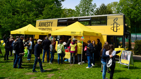 Das Bild zeigt einen gelben Doppeldecker-Bus im Hintergrund, viele Menschen, manche mit Amnesty-Leibchen