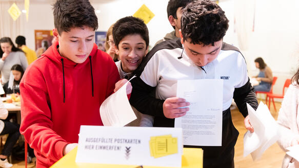 Das Bild zeigt mehrere Jugendliche, die Briefe in einen gelben Kasten werfen
