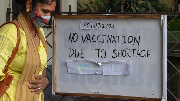 Das Bild zeigt eine Frau rechts im Bild, die an einem Schild vorbeiläuft mit der Nachricht "Vaccine Shortage"