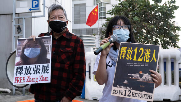 Das Bild zeigt zwei Menschen, die Protestschilder hochalten, sie stehen vor einem Gebäude mit chinesischer Flagge, auf einem Plakat ist das Foto einer Frau zu sehen sowie der Schriftzug Zhang Zhan