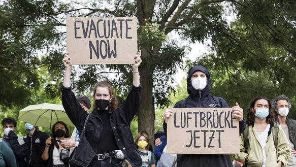 Das Bild zeigt eine Demonstration mit mehreren Menschen, zwei halten Schilder mit der Aufschrift "Evacuate Now", "Luftbrücke Jetzt"