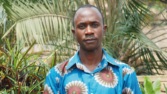 Ein junger Mann in einem kurzärmeligen Hemd steht vor einem Palmenhain und blickt in die Kamera.