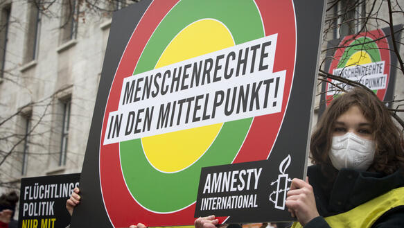 Das Bild zeigt eine Protestaktion bei der eine Maske tragende Frau im Vordergrund steht und ein Amnesty-Schild mit der Aufschrift "Menschenrechte in den Mittelpunkt!" hält.