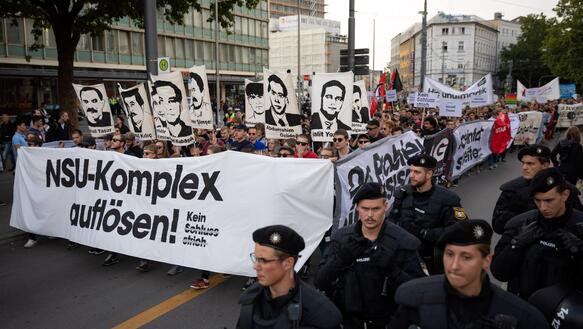 Das Bild zeigt eine Demonstration, im Vordergrund Polizeikräfte, dahinter Demonstrierende mit einem Banner "NSU-Komplex auflösen