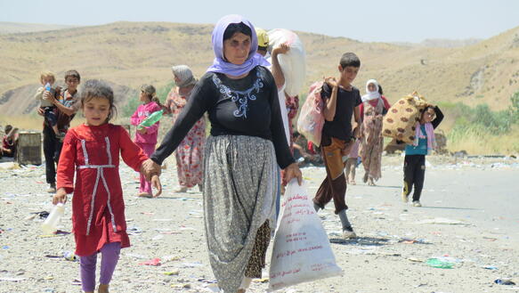 Das Bild zeigt eine Frau mit Kind im Vordergrund, dahinter viele weitere Kinder, sie gehen mit viel Gepäck eine Straße entlang