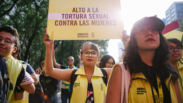 Das Bild zeigt eine Demonstrantin zwischen weiteren Demonstrierenden mit gelben Amensty-Westen, die ein spanisches Schild hält, das zur Beendigung sexueller Gewalt gegen Frauen aufruft.