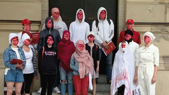 Menschen mit rotweißen Gesichtsmasken stehen als Gruppe zusammen auf einer Treppe vor einem Gebäude.