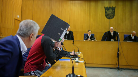 Gerichtssaal, in dem Richter sitzen und ein Angeklagter, der einen Aktenordner hochält, um seinen Kopf zu verbergen.