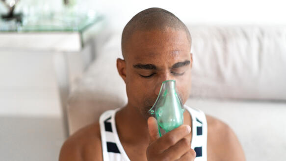 Das Foto zeigt einen PoC-Mann mit geschlossenen Augen, der sich ein Inhaliergerät über Nase und Mund hält.