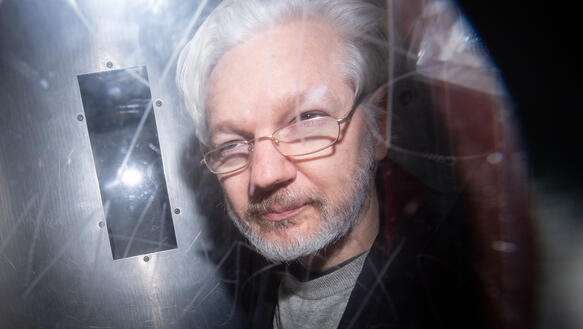 Aufnahme von Julian Assange durch eine zerkratze Scheibe hindurch. Er trägt eine Brille.