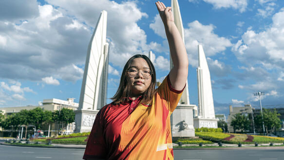 Eine junge thailändische Aktivistin steht vor einem Denkmal und hebt ihre linke Hand in den Himmel, über den Wolken treiben.