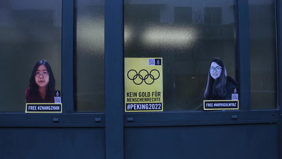 In den Fenstern einer Eingangstür sind zwei ausgeschnitte Bilder von zwei Frauen und ein gelbes kleines Plakat, auf dem die fünf olympischen Ringe zu sehen und die Aufschrift "Kein Gold für Menschenrechte #Peking2022"