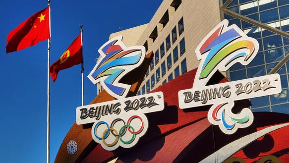 Das Bild zeigt ein Werbeschild der Olympischen Spiele vor einem Gebäude: Man sieht die Olympischen Ringe und den Schriftzug "Beijing". Dahinter sind chinesische Nationalflaggen zu sehen.