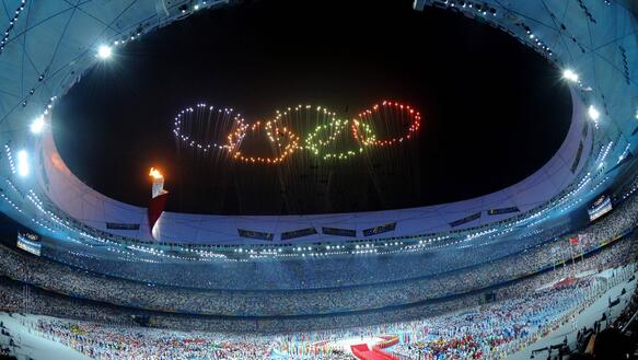 Ein Sportstadium bei Nacht, in dem viele Menschen sind. Mit Lichteffekten sind im Nachthimmel die fünf olympischen Ringe zu sehen. 