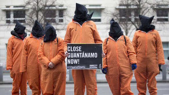 Das Bild zeigt sechs Menschen in orangenen Overalls mit schwarzen Säcken über den Köpfen. Eine Person hält ein Schild mit der Aufschrift "Close Guantanamo now!".