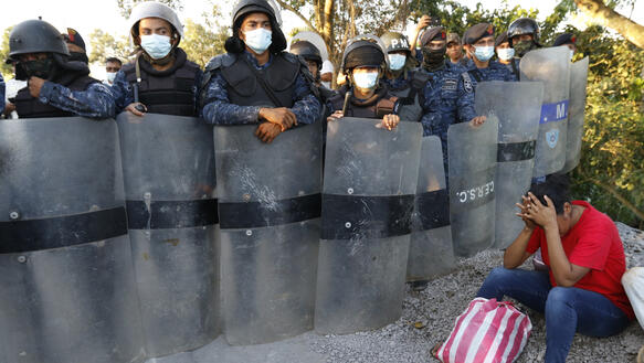 Sicherheitskräfte mit Helmen, Schilden und Mundnasenschutzmasken bilden mit ihren Schilden eine Barriere, vor der eine Frau sitzt, die ihr Gesicht in ihren Händen vergräbt.