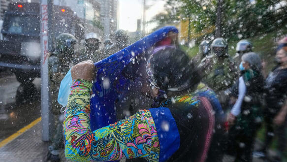 Eine Frau steht vor einer Gruppe von Polizisten mit Helmen und hält ein Tuch in die Höhe, um sich vor spritzendem Wasser zu schützen.