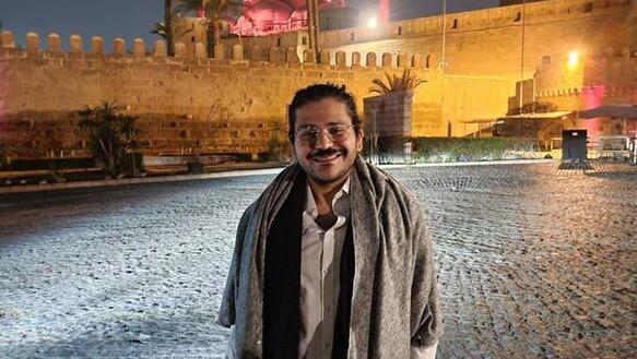 Patrick George Zaki steht nachts auf einem Platz, im Hintergrund steht eine hell erleuchtete Moschee.