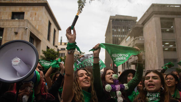 Das Bild zeigt eine Menschenmenge, darunter viele Frauen, die lachen, grüne Banner in die Luft halten. Eine Frau hält ein Megaphon. 