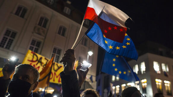 Das Bild zeigt eine Demonstration bei Nacht, die Silhouette eines Mannes, der zwei Flaggen in die Höhe hält - die EU-Flagge und die polnische Flagge