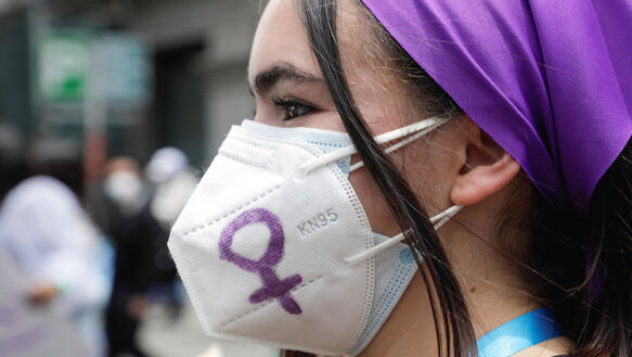 Eine junge Frau trägt ein Kopftuch und eine Mundnasenschutzmaske, auf die ein Weiblichkeitssymbol gemalt ist.