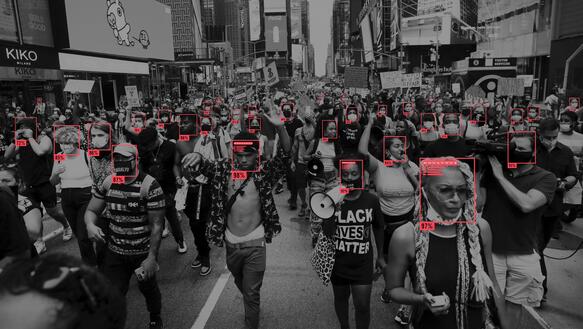 Das Bild zeigt eine Demonstration mit vielen Menschen. Auf den Gesichtern mancher Demonstrierender sind rote Vierrecke zu sehen.
