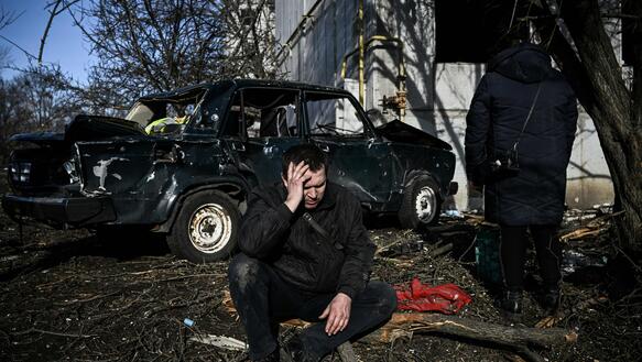 Das Bild zeigt einen Mann der am Boden kniet und seinen Kopf mit der Hand stützt, im Hintergrund sieht man ein zerstörtes Auto und Gebäude.