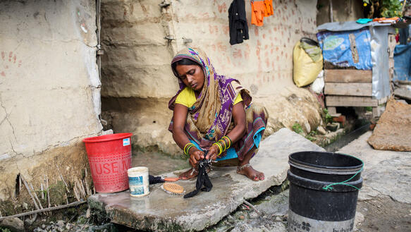Eine Frau hockt vor einem steinernen Haus und wäscht Wäsche, um sie herum stehen Eimer.