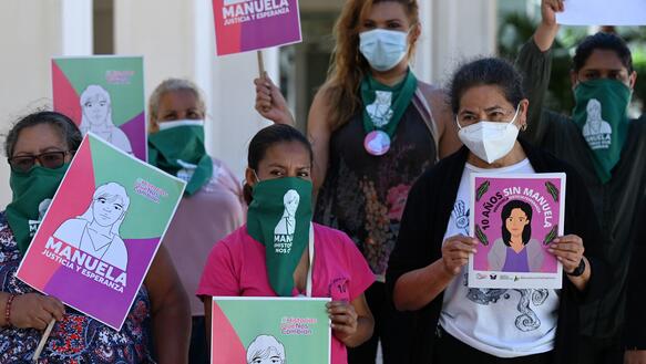 Das Foto zeigt eine Gruppe von sechs Frauen mit Mundschutz, die Schilder mit dem gezeichneten Porträt einer Frau hochhalten. Auf den Schildern steht unter anderem: Manuela.