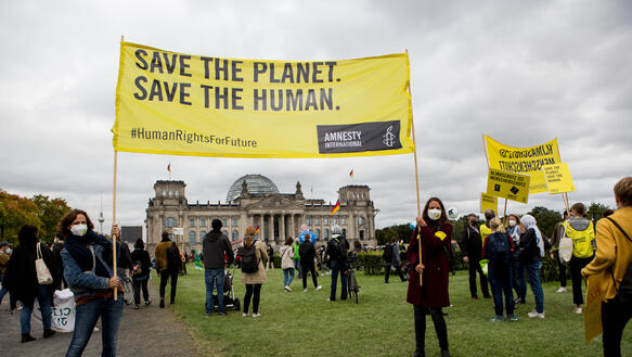 Zwei Personen halten ein Transparent mit der Aufschrift "Save the Planet. Save the Human" hoch. Im Hintergrund ist das Reichstagsgebäude in Berlin zu sehen sowie viele weitere Menschen. 