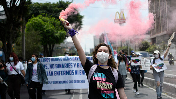 Das Bild zeigt einen Protestzug, im Vordergrund eine junge Frau, die eine Rauchfackel mit rosa Rauch in der Hand hält 
