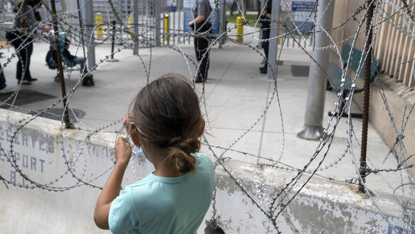 Ein Mädchen steht vor einem Stacheldrahtzaun hinter dem sich Sicherheitskräfte und eine Frau mit Kinderwagen befinden.