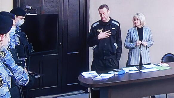 Alexej Nawalny steht neben seiner Anwältin hinter einem Tisch und legt seine rechte Hand auf seine Brust. Neben ihm stehend zwei unformierte Justizbeamte mit Mundschutz.