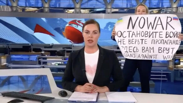 Das Bild zeigt eine TV-Nachrichtensprecherin hinter der eine Frau ein Schild hochhält, auf dem unter anderem "No War" steht.