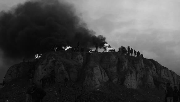 Auf einem Felsen stehen Menschen, links ihnen brennt etwas, von diesem Feuer aus steigt dichter schwarzer Rauch auf.