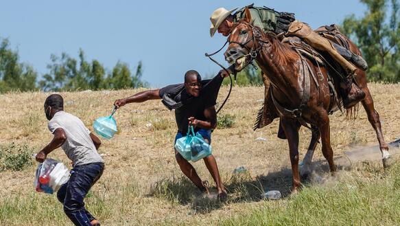 Auf einer Wiese versucht ein Mann auf einem Pferd mit Lasso und Hut einen Mann am T-Shirt festzuhalten, der versucht zu entkommen. Eine weitere Person läuft daneben.