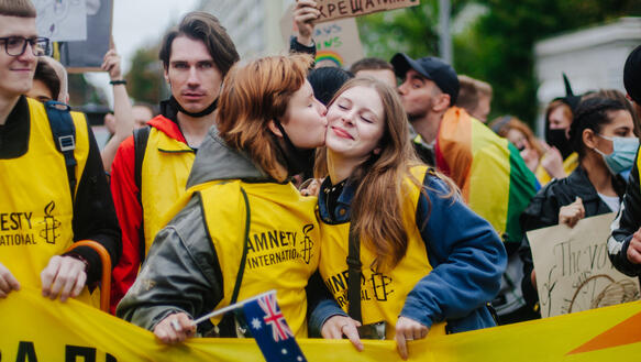 Das Foto zeigt eine Gruppe junger Menschen, die Leibchen mit dem Amnesty-Logo tragen. Sie stehen inmitten einer großen Menschenmenge. In der Bildmitte küsste eine junge Frau eine andere junge Frau auf die rechte Wange.